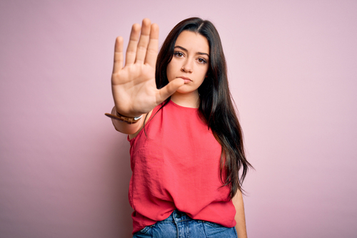 A young woman with long dark hair wearing a red blouse extends her hand forward in a stop gesture, with a serious expression on her face.