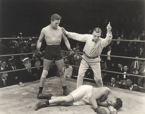 A vintage black and white photo of a boxing match. One boxer stands victorious, his arm raised by the referee, while the other lies defeated on the canvas. The crowd watches intently in the background.